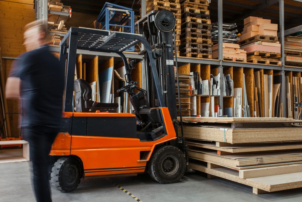 Warehouse with an orange forklift parked next to stacks of wood boards. Shelves in the background hold more wood and materials, and a person is walking by, slightly blurred.
